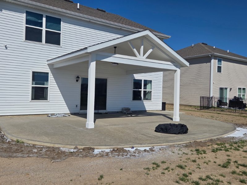 Covered porch with white gable roof and decorative truss over a large curved concrete patio on a white sided home